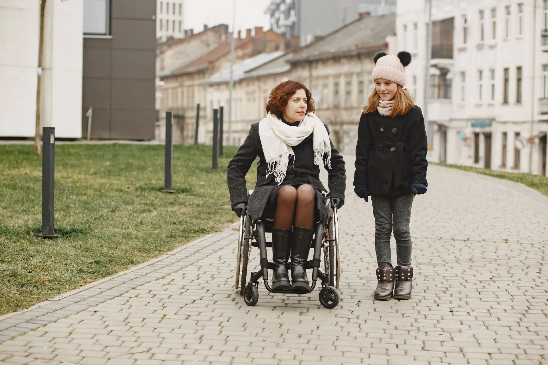 A woman in a wheelchair and a girl walking on a cobbled path in urban setting