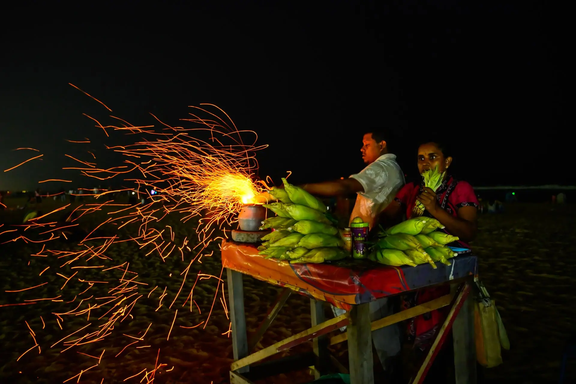 Dynamic nighttime scene of a street vendor grilling corn on Chennai beach, sparks flying
