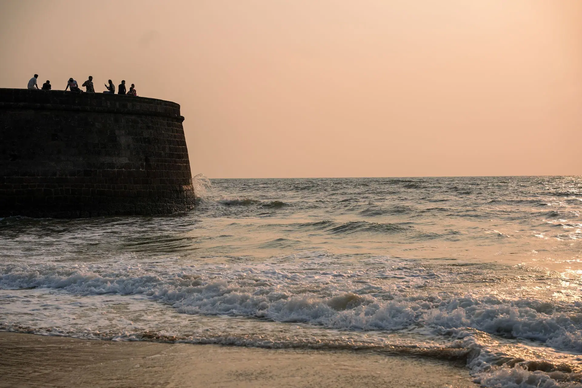 Silhouetted figures on a historic fort wall during a peaceful Goa sunset by the sea