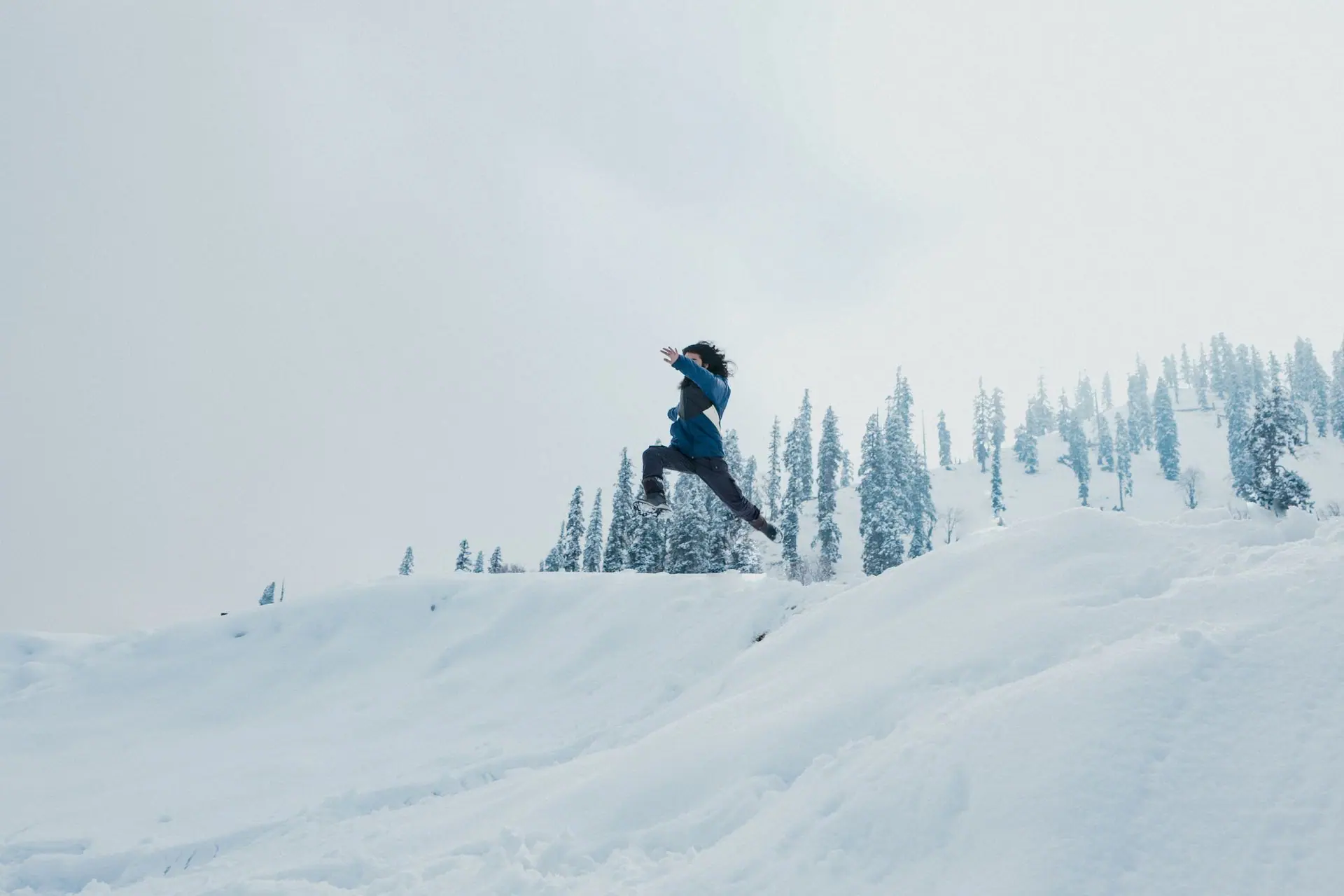A man leaps across a snow-covered hillside against a backdrop of trees in Gulmarg