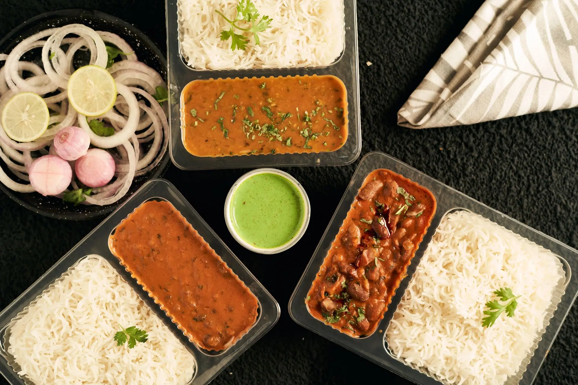 Top view of Indian curry, rice, and greens in takeaway containers with sliced onions