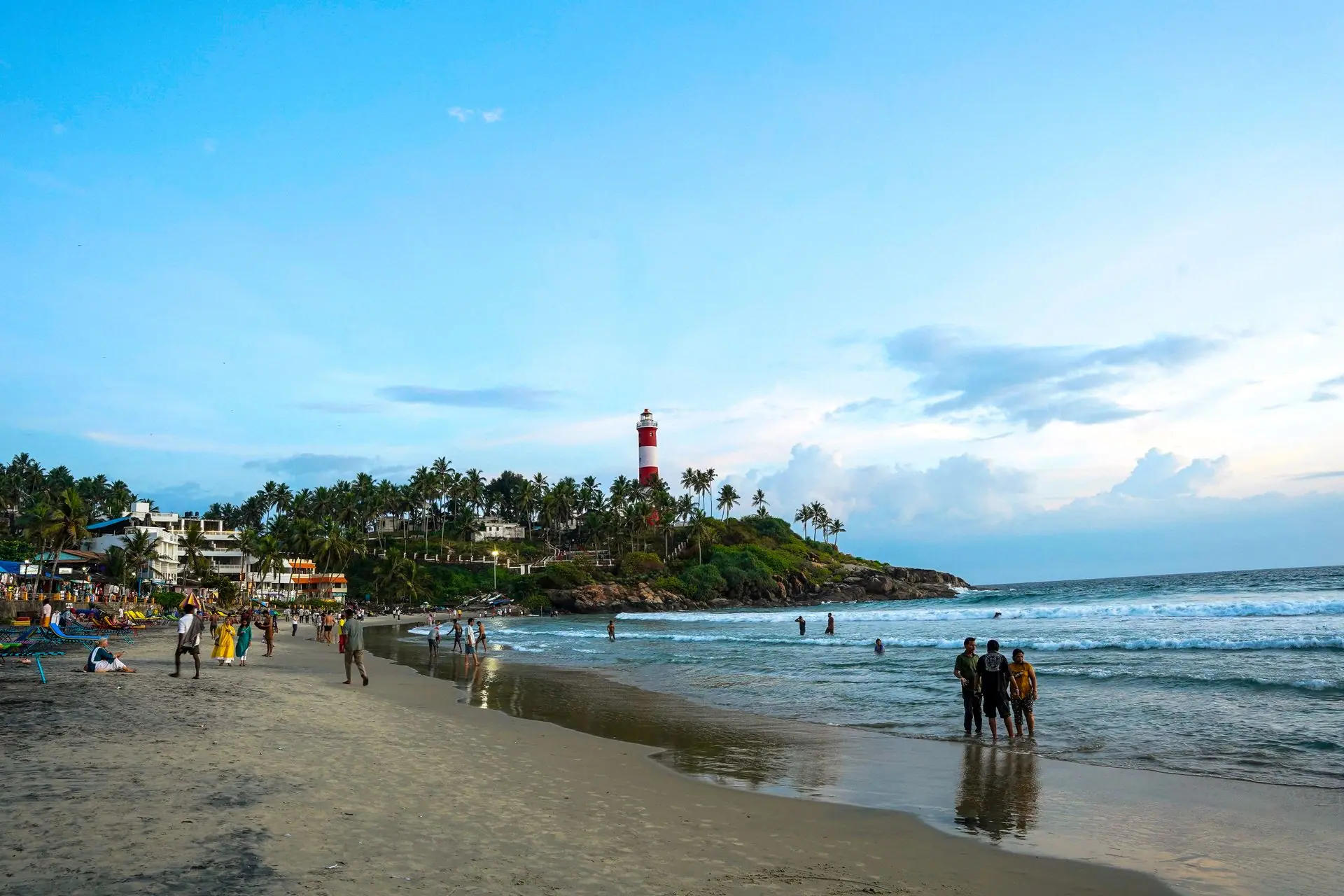 Kovalam Beach scene with lighthouse, palm trees, and people enjoying a blue sky day