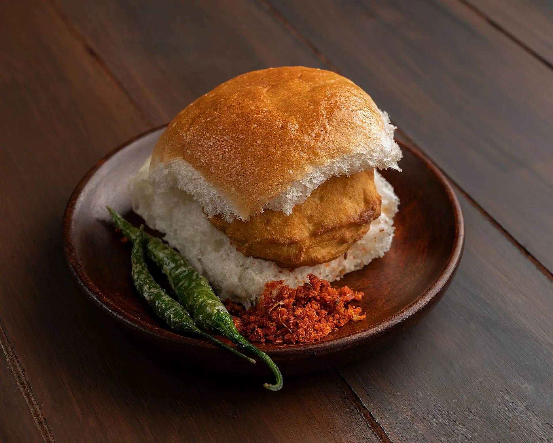 A mouthwatering Vada Pav served with green chili and chutney on a wooden plate