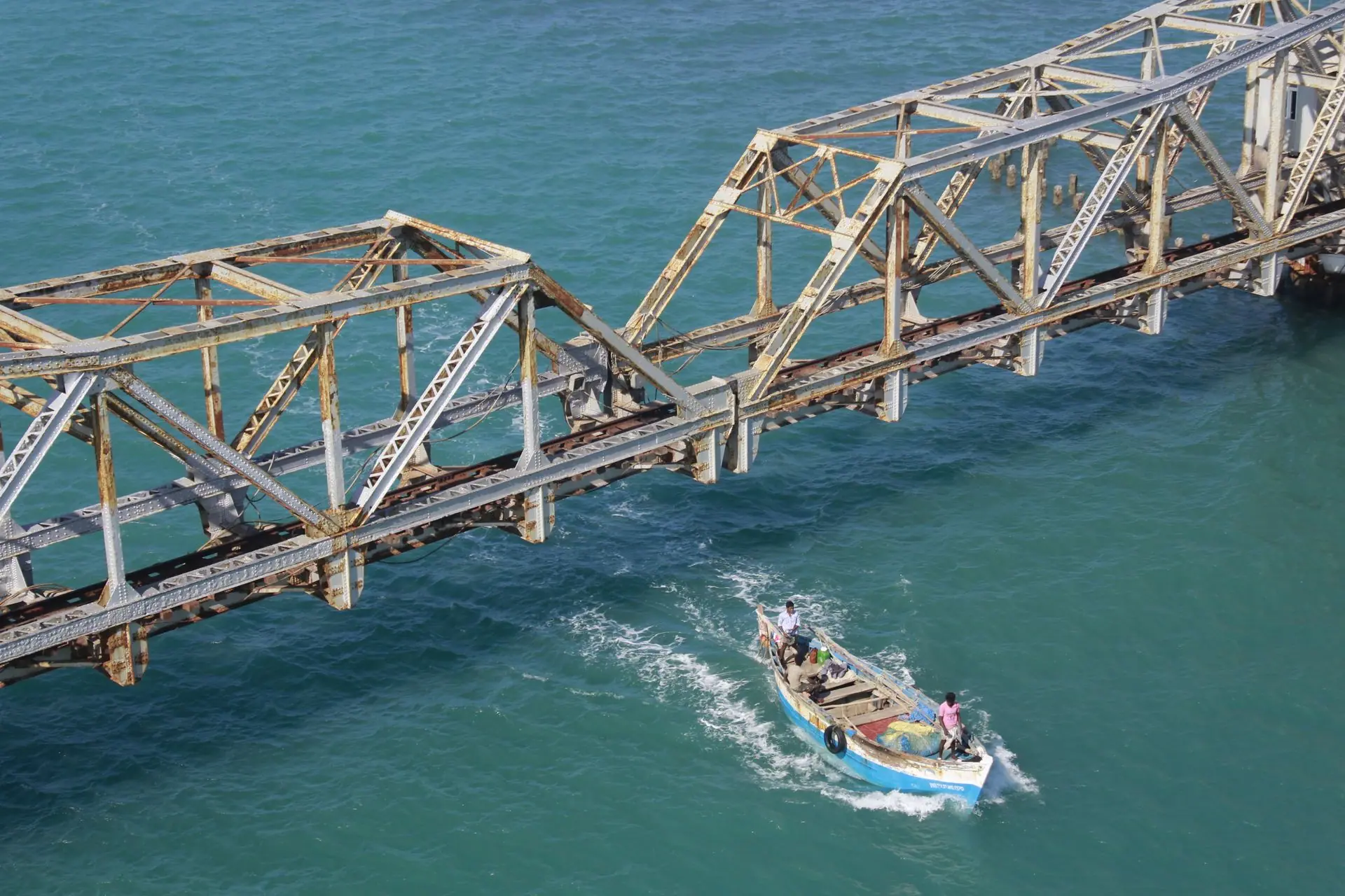 A fishing boat navigates beneath the historic rusty Pamban Bridge in clear blue waters