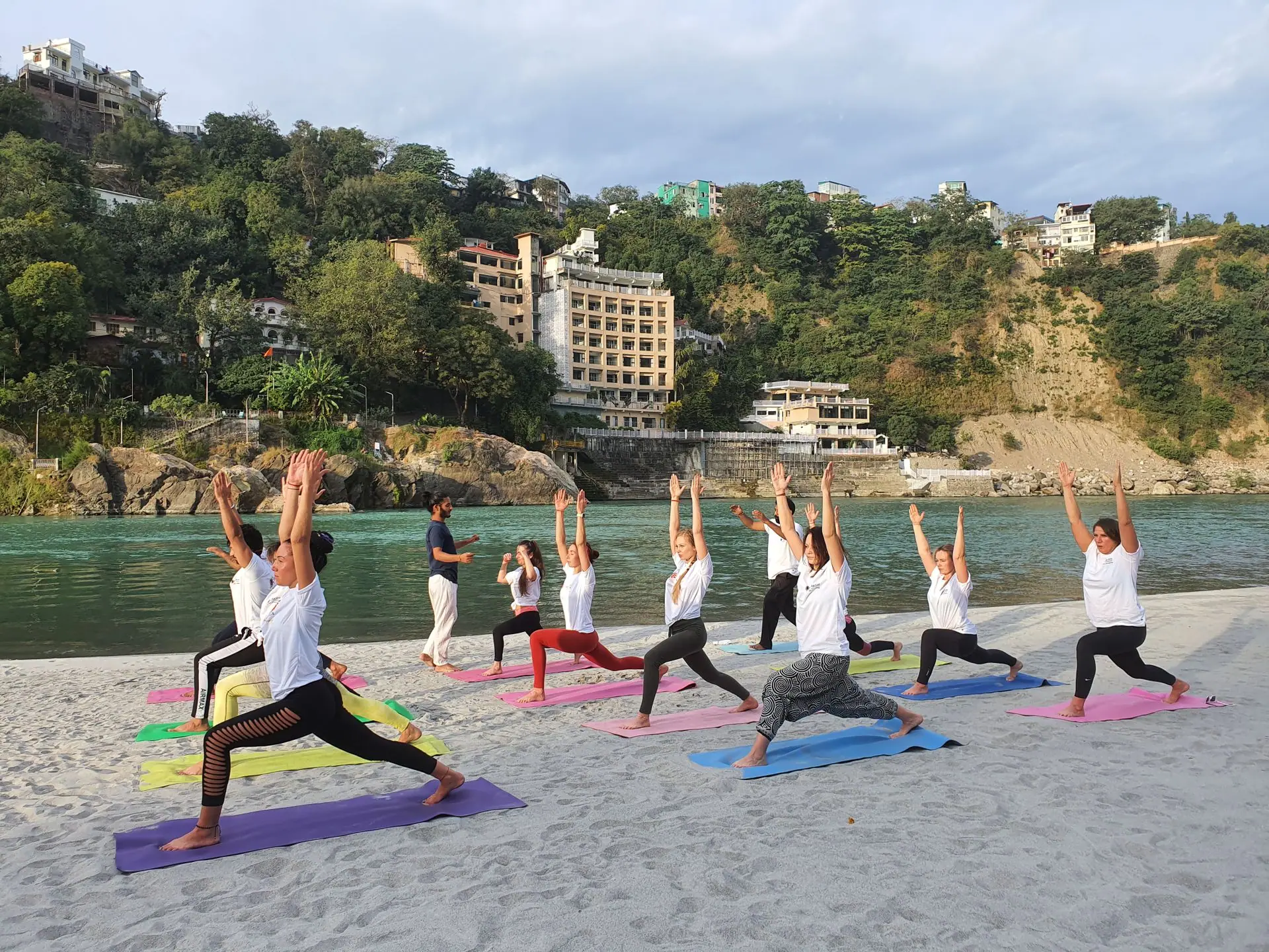 A group of adults practicing yoga on mats by a river with scenic hills in Rishikesh