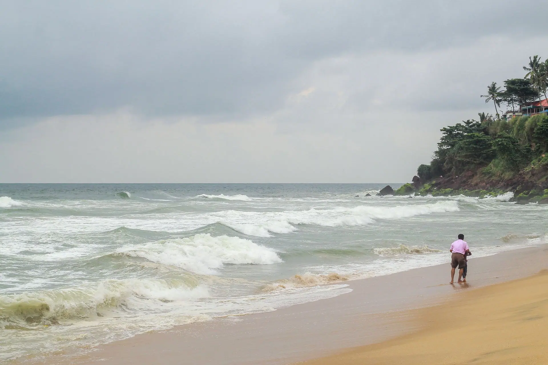 A father and child walk along the sandy shores of Varkala Beach, India, by the ocean waves