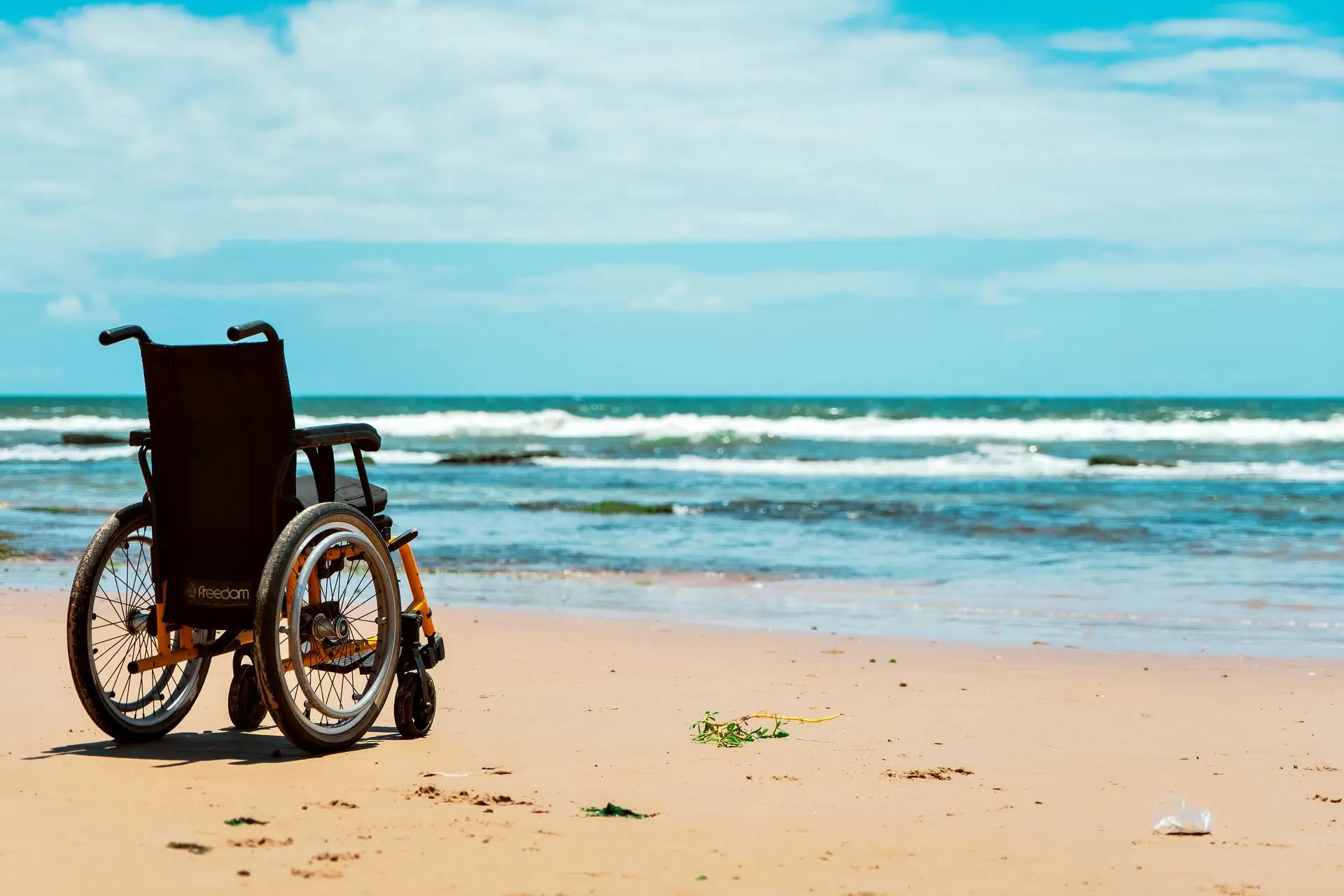 A wheelchair sits on a serene beach, facing the calming sea waves under a clear sky