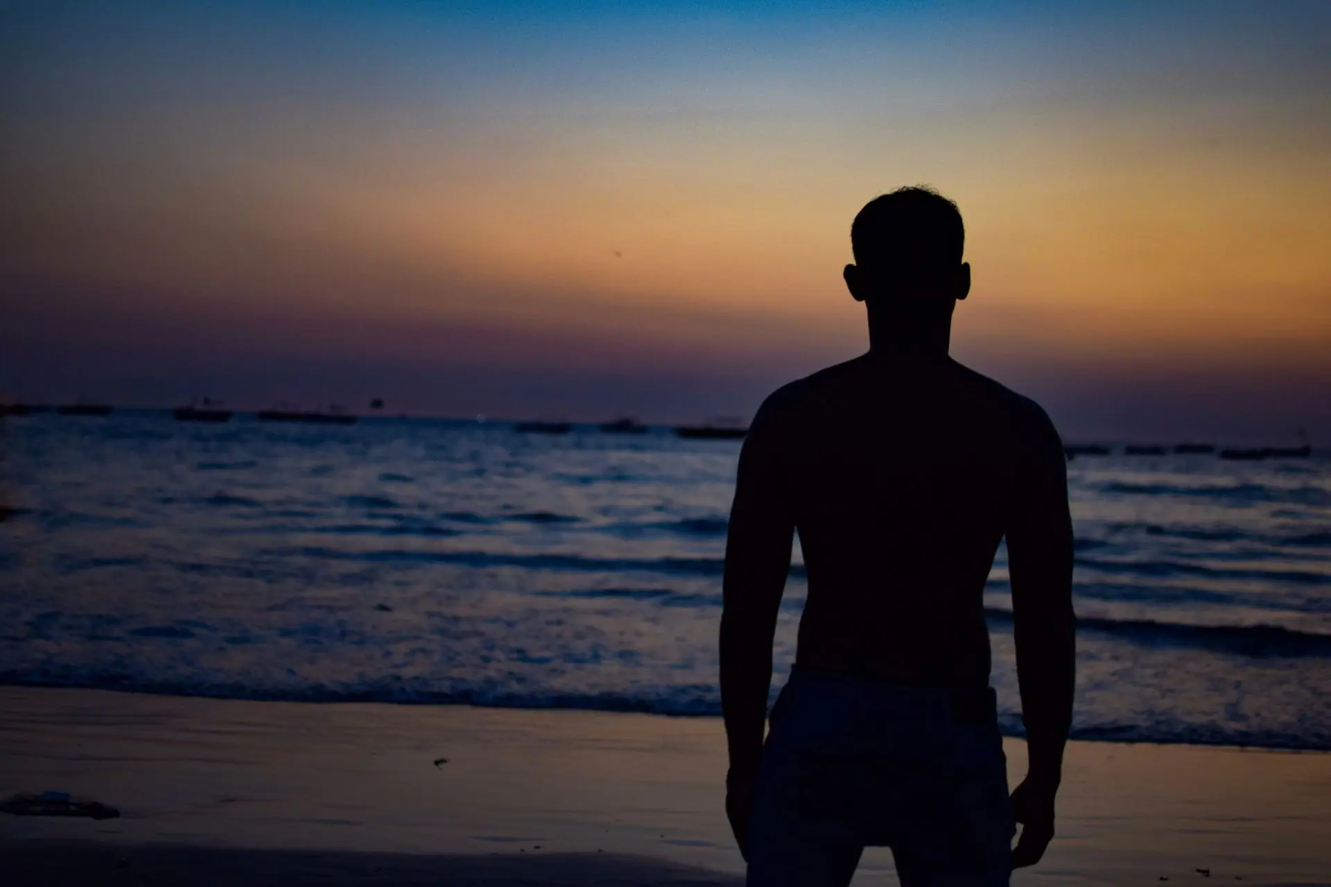 A man stands on the shore of a Goa beach, silhouetted against a vibrant sunset