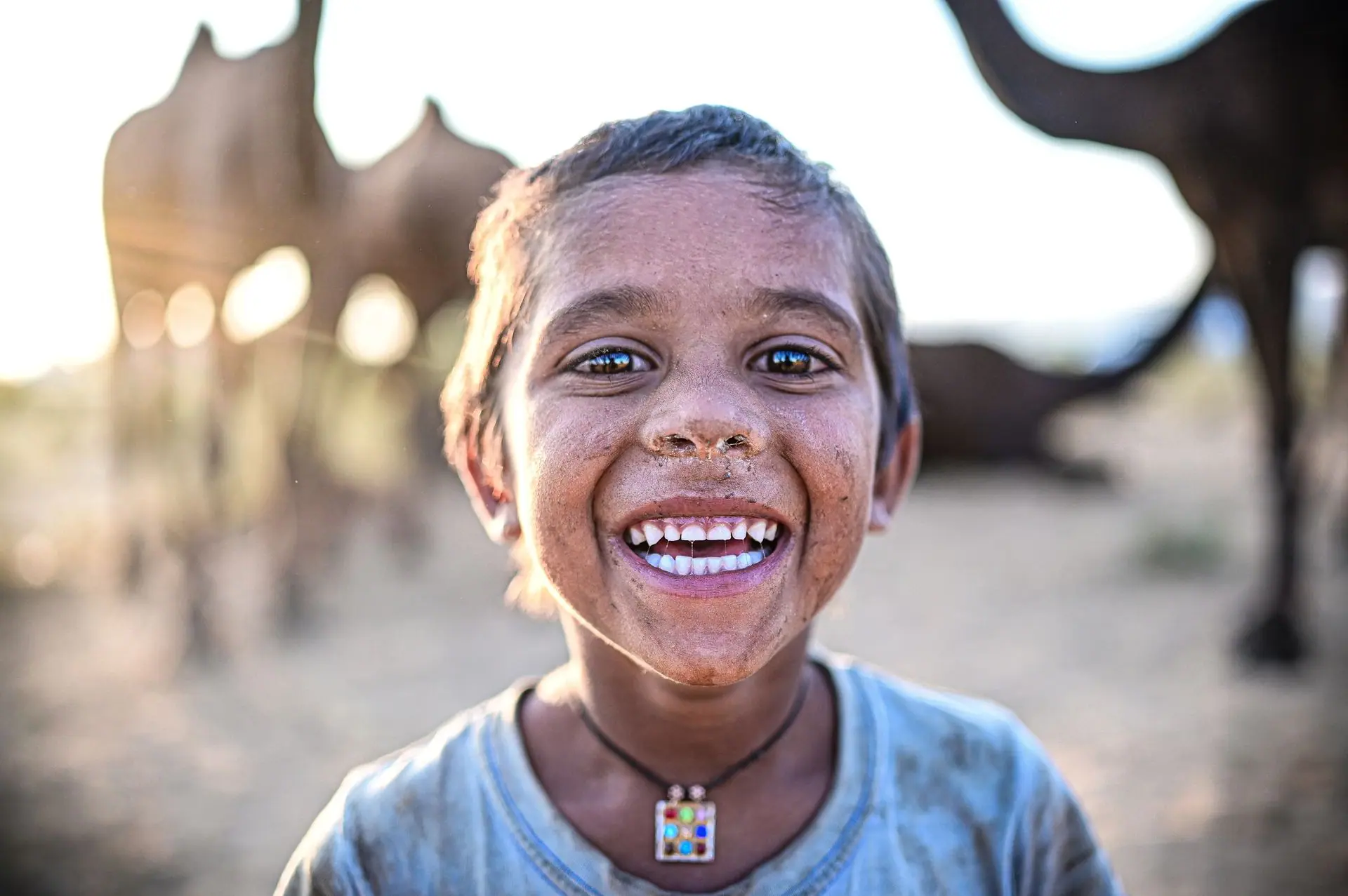A cheerful child with a dirty face smiles widely in the Pushkar desert, surrounded by camels and warm light