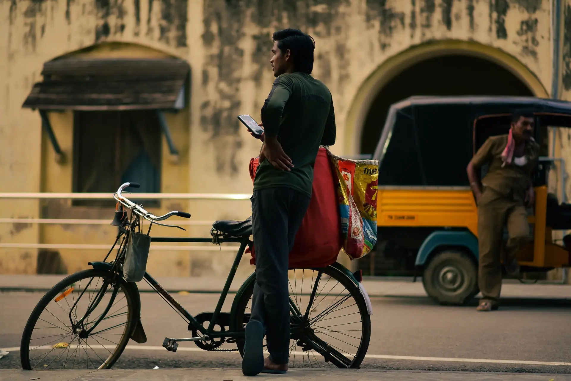 A man stands by his bicycle on a street, with an auto rickshaw nearby, against a rustic backdrop