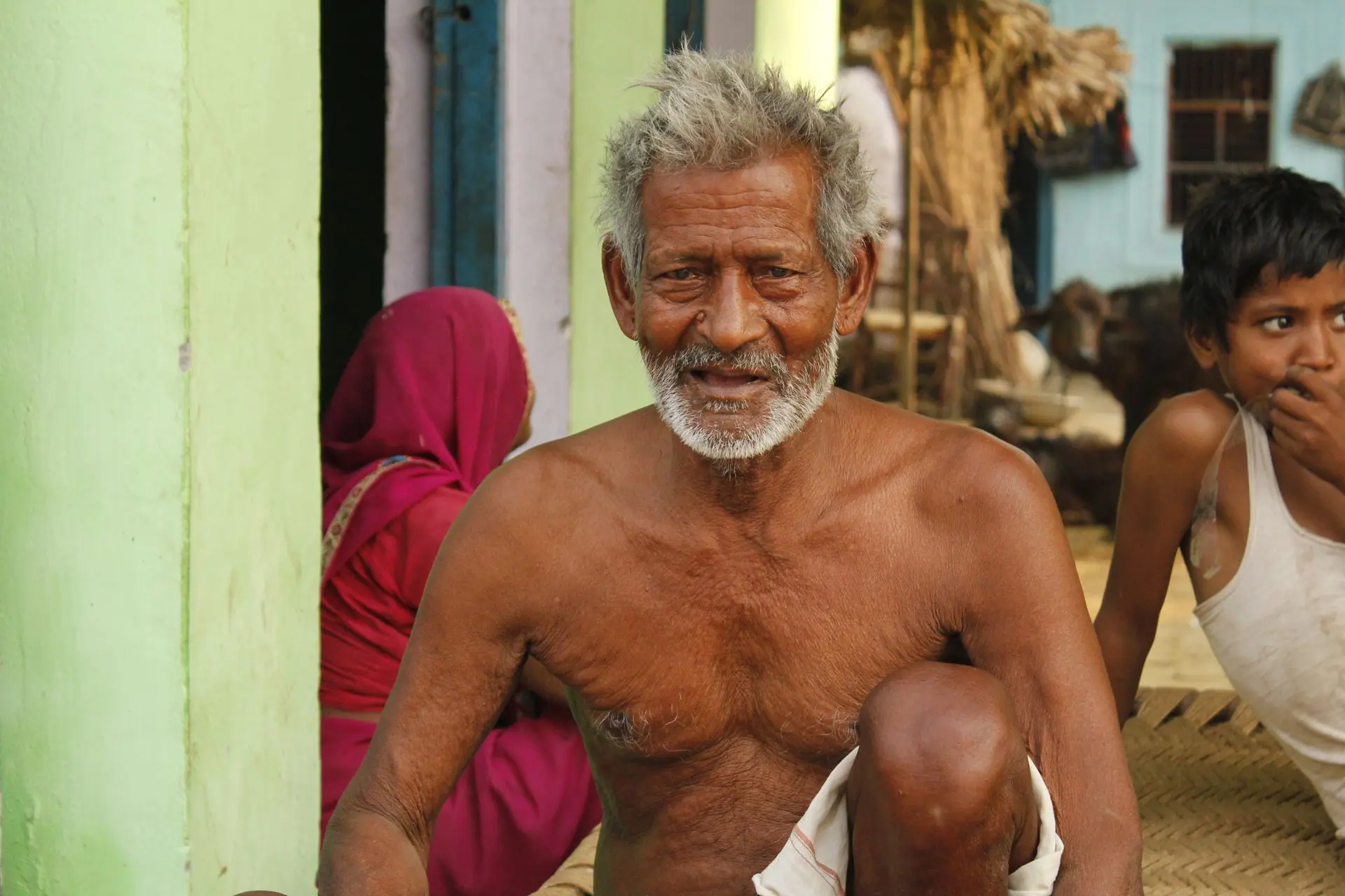 A bearded senior man with white hair sits outdoors in a colorful Indian village