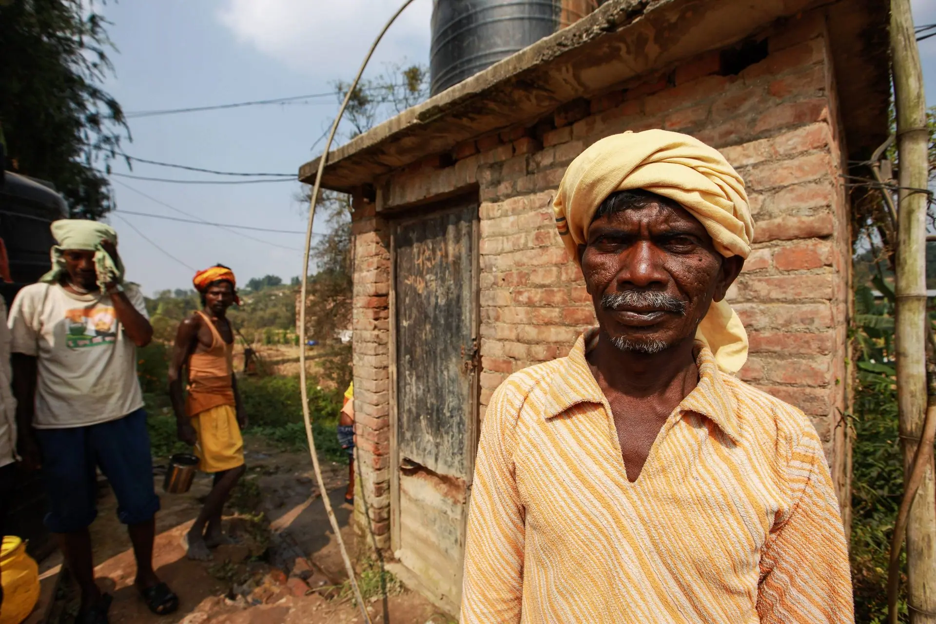 Elderly man in rural India wearing a turban, standing by a brick structure
